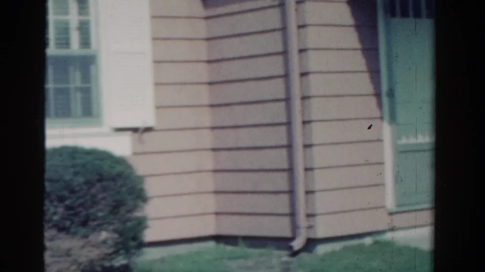 1958 Ontario residential exterior with clapboard siding, white shutters, downspout, green door edge, lawn, and shrubbery.