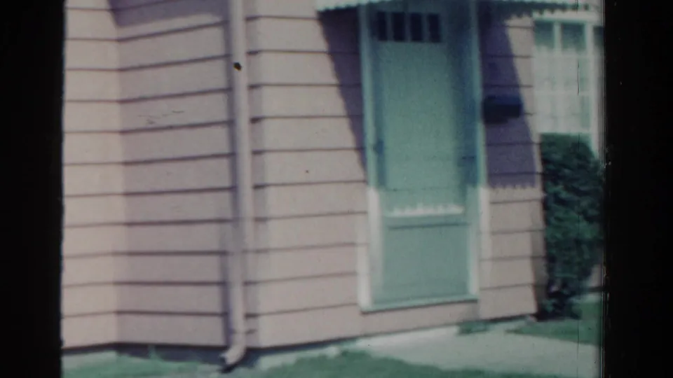 1958 Ontario home-movie frame showing a beige-sided house exterior with a mint-green door, white window trim, shrubs, and lawn.