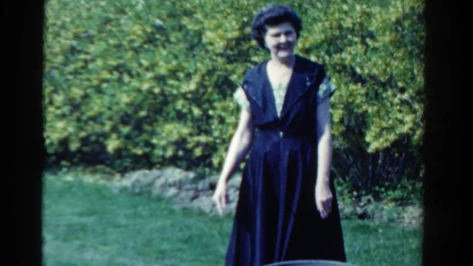 1958 Ontario home-movie frame of a woman in a dark sleeveless dress smiling and waving from a green garden lawn.