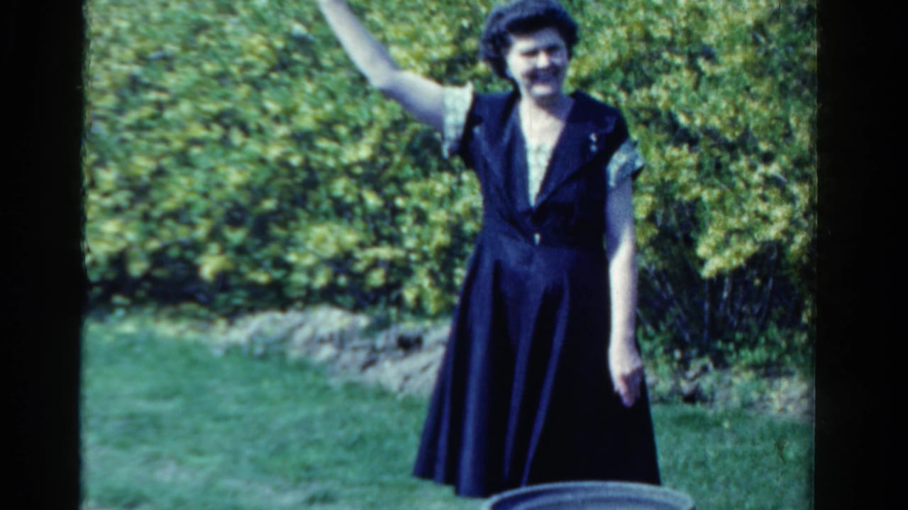 Woman in a dark dress waves while standing on a lawn in front of a hedge.