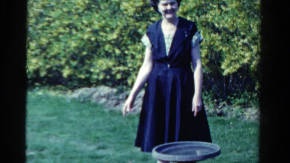 A woman in a dark 1950s-style dress stands on a green lawn near a birdbath in Ontario home-movie footage.