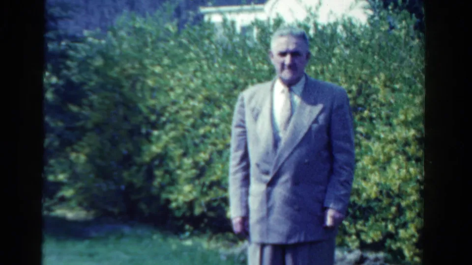 An older man in a light suit stands in the same sunny Ontario garden, facing the home-movie camera.