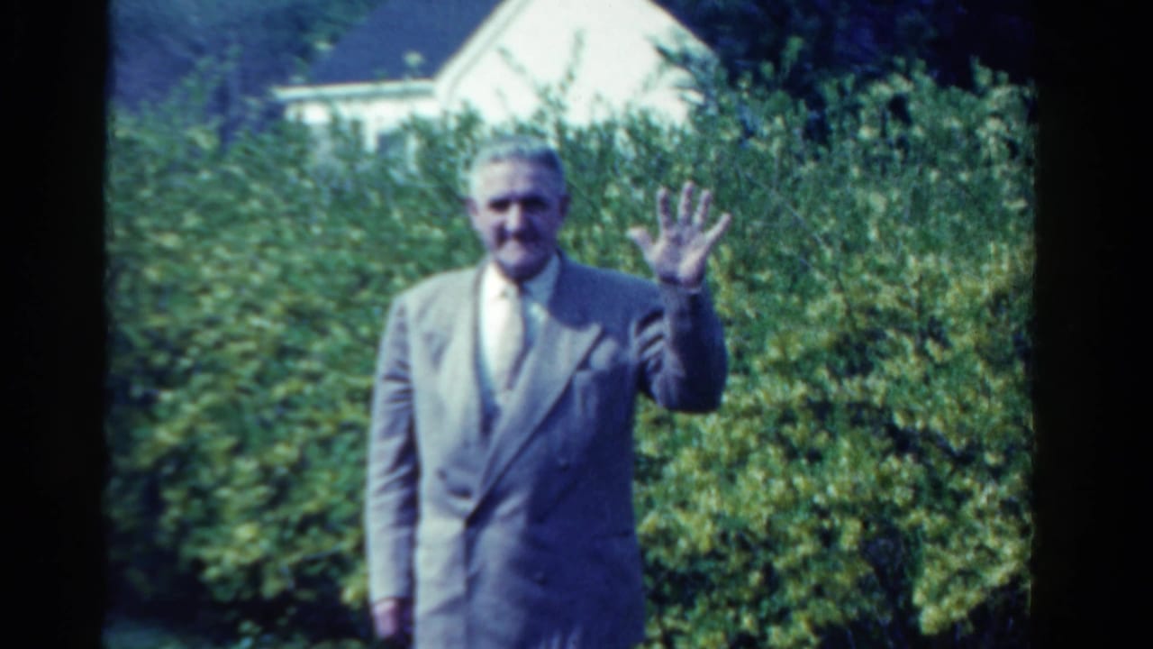 In the Ontario garden scene, the suited adult man faces the camera and raises his hand in a brief wave.