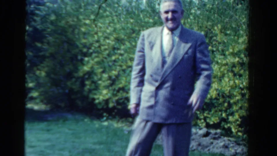 The suited man appears closer in frame, standing casually in the garden with green bushes and lawn behind him.