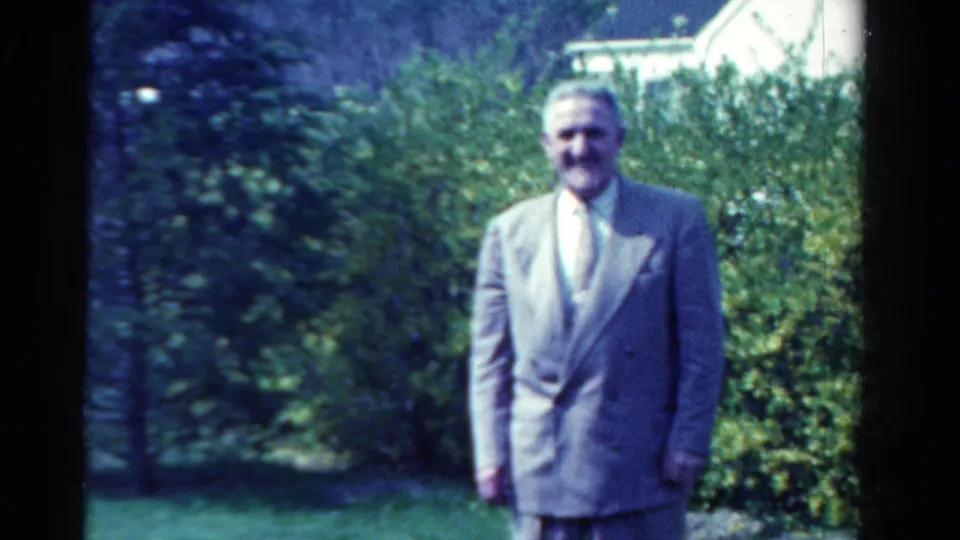 The adult man remains facing the camera in the garden, with a light-colored roofline partly visible beyond the hedge.