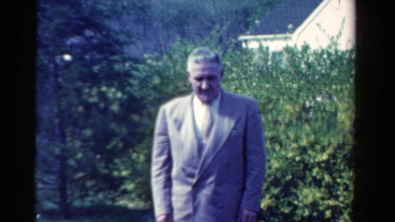 Man in a light-colored suit stands outdoors near shrubs and a sunlit garden.