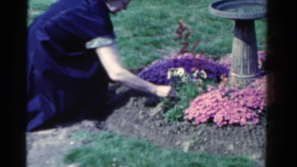 The kneeling woman tends a flowerbed in the Ontario garden, with bright blooms and a stone birdbath visible nearby.