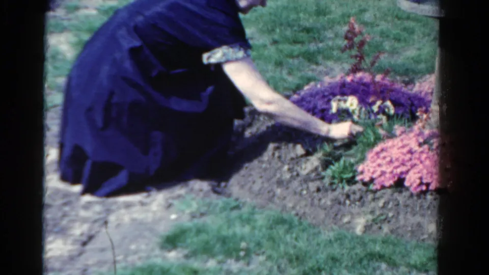 The 8mm frame shows a woman kneeling low beside a memorial-style flowerbed, her hand near the plants and soil.