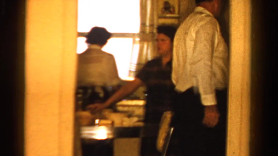 1958 Ontario home-movie frame looking through a doorway toward adults gathered in a warmly lit kitchen.