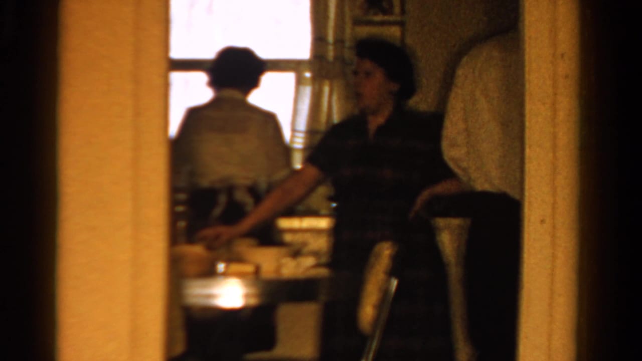1958 Ontario home-movie frame of a woman in a dark dress near a kitchen counter, with another adult seen from behind at a window.