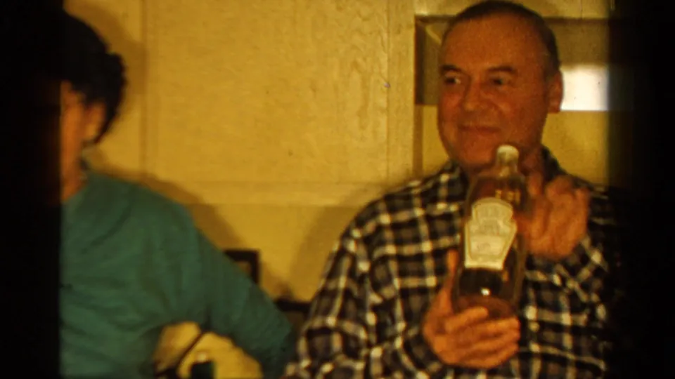 1958 Ontario home-movie frame showing a man in a plaid shirt holding a glass condiment bottle in a kitchen interior.