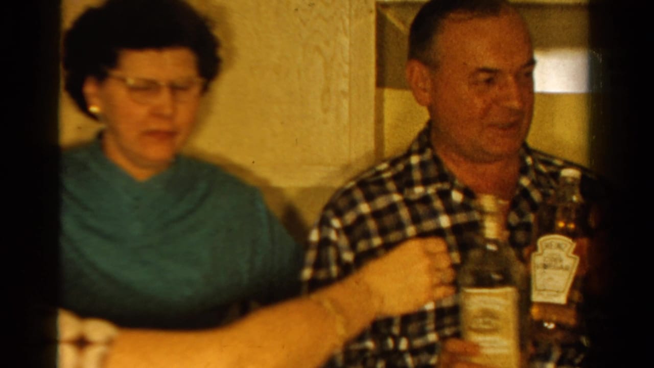1958 Ontario home-movie frame showing adults in a warm kitchen as a man in a checkered shirt holds bottles near a seated woman in teal.