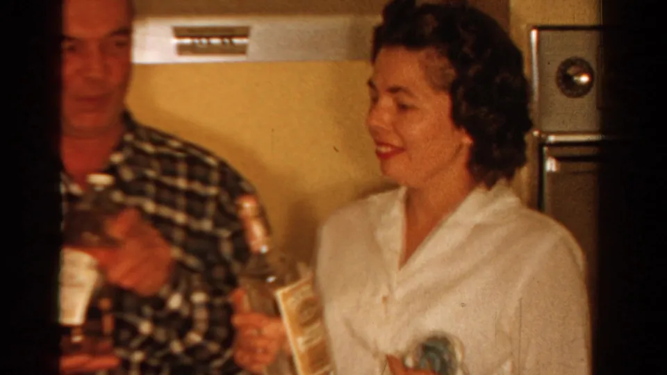 1958 Ontario home-movie frame showing a smiling woman in a white blouse holding a clear bottle in a private kitchen interior.