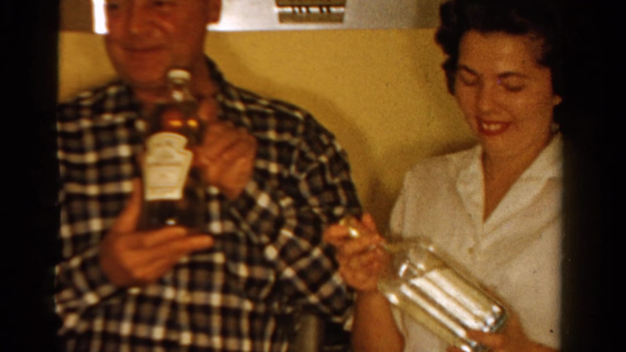 1958 Ontario home-movie frame of adults seated indoors, with a man holding a bottle and a woman in a white blouse smiling with a glass.