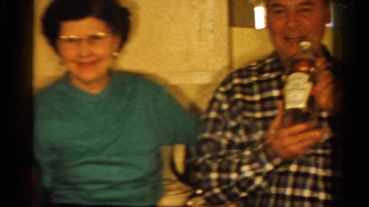 1958 Ontario home-movie frame of a woman in a green sweater seated near a man in plaid holding a labeled bottle indoors.