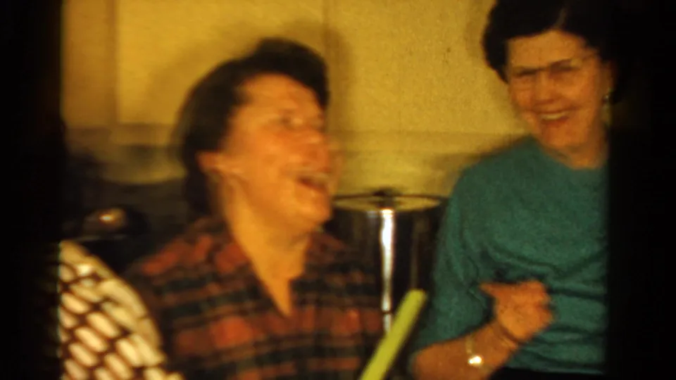 1958 Ontario home-movie frame of women laughing in a private interior, one in a plaid shirt and another in a green sweater.