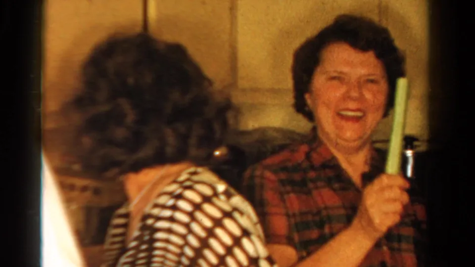 1958 Ontario home-movie frame of a laughing woman in a red plaid shirt holding a green candlestick in a domestic interior.