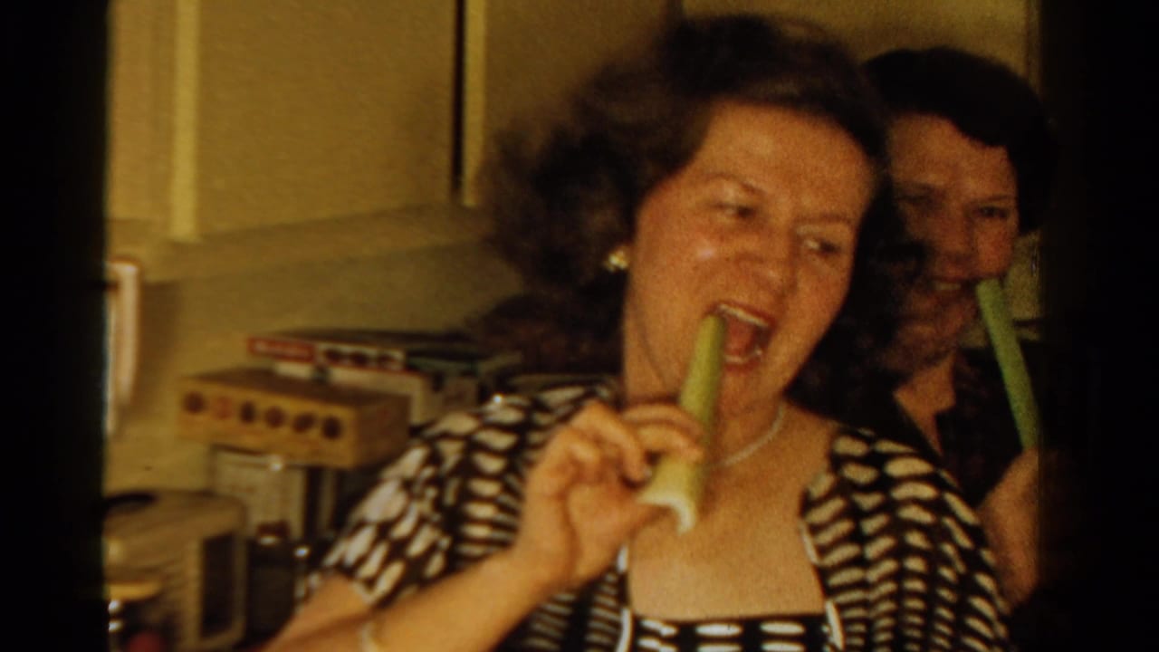 A woman bites a celery stick while another person stands partly behind her in a kitchen.