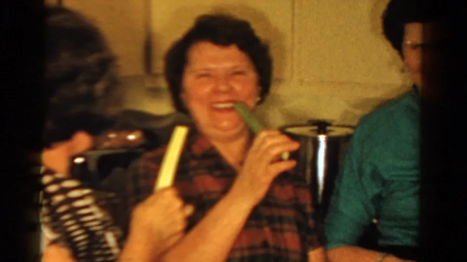 A woman in a red plaid shirt laughs while holding celery in an Ontario kitchen, with other adults partly visible around her.