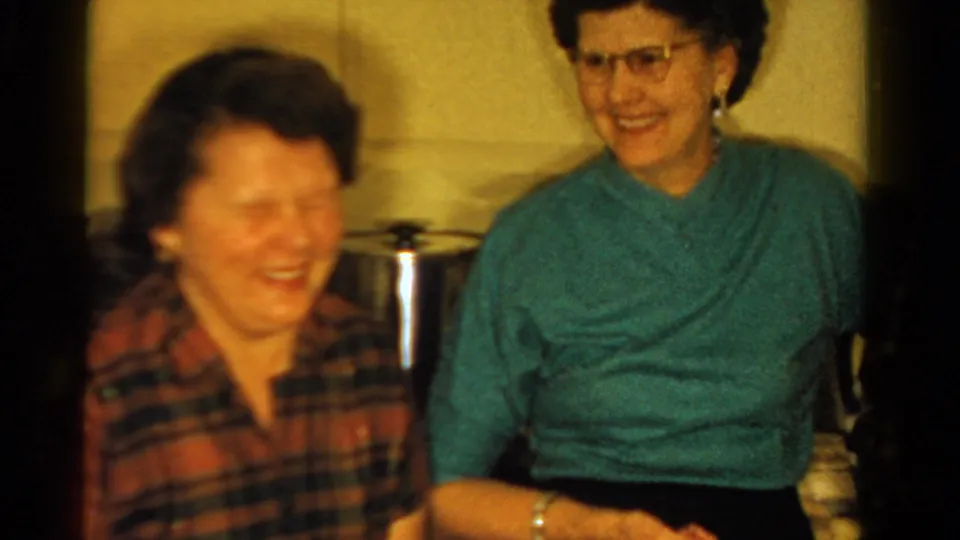 Two adult women laugh in a warmly lit Ontario kitchen, with cookware and cabinets visible behind them.