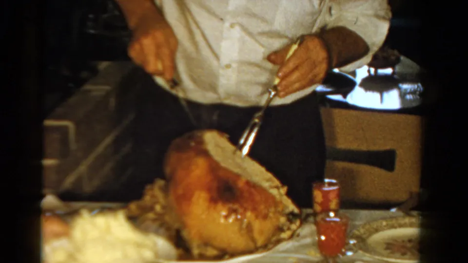A 1958 home-movie frame shows an adult leaning over a roast turkey with carving utensils during a household meal.