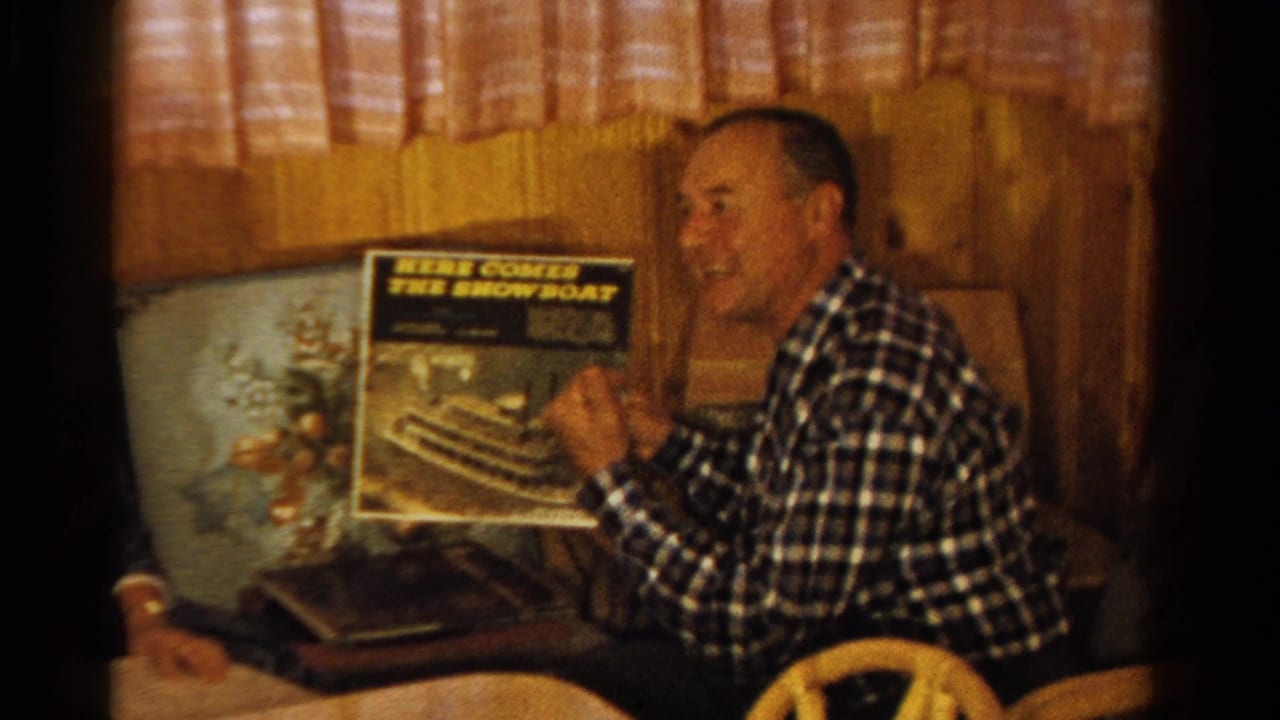 A seated man in a plaid shirt smiles while holding a record album in a wood-paneled Ontario interior.