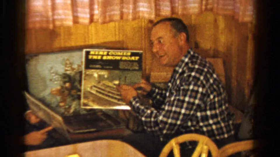 The seated man continues smiling with the record sleeve visible as another person sits partly in frame.