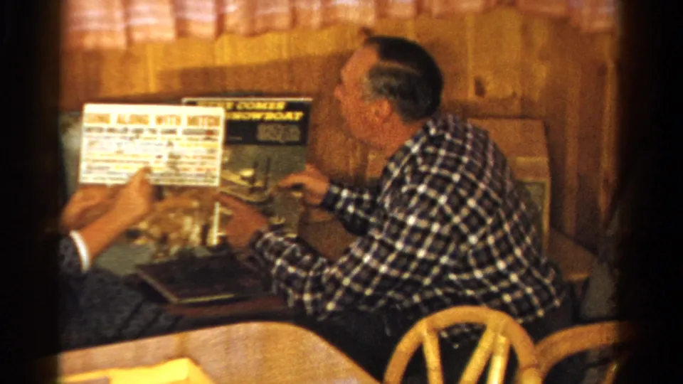 A seated man in plaid turns toward an off-camera companion holding papers, with the record album still visible nearby.
