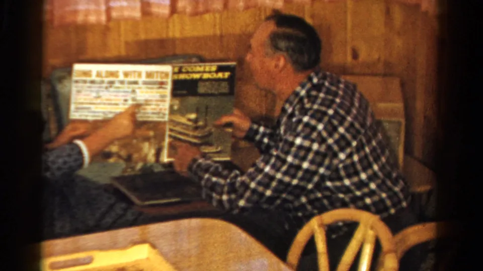 In a 1958 Ontario home interior, a seated man and companion examine record album covers in a wood-paneled room.