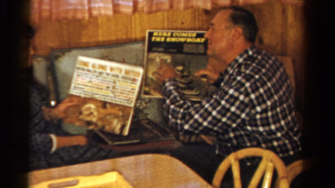 A man in a plaid shirt holds up an album as another person shows a record cover inside a private Ontario home setting.