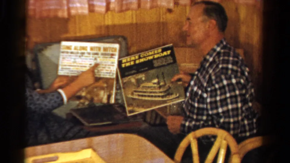 The companion points at text on a record cover as the seated man looks toward the albums in the 1958 Ontario home-movie scene.