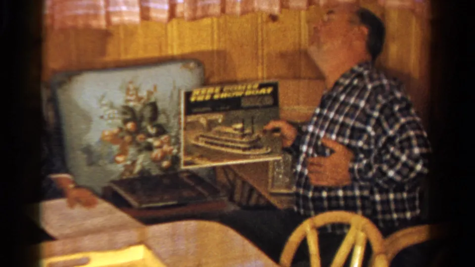 The seated man reacts with a smile or laugh while holding an album cover in the warm 8mm Ontario interior.