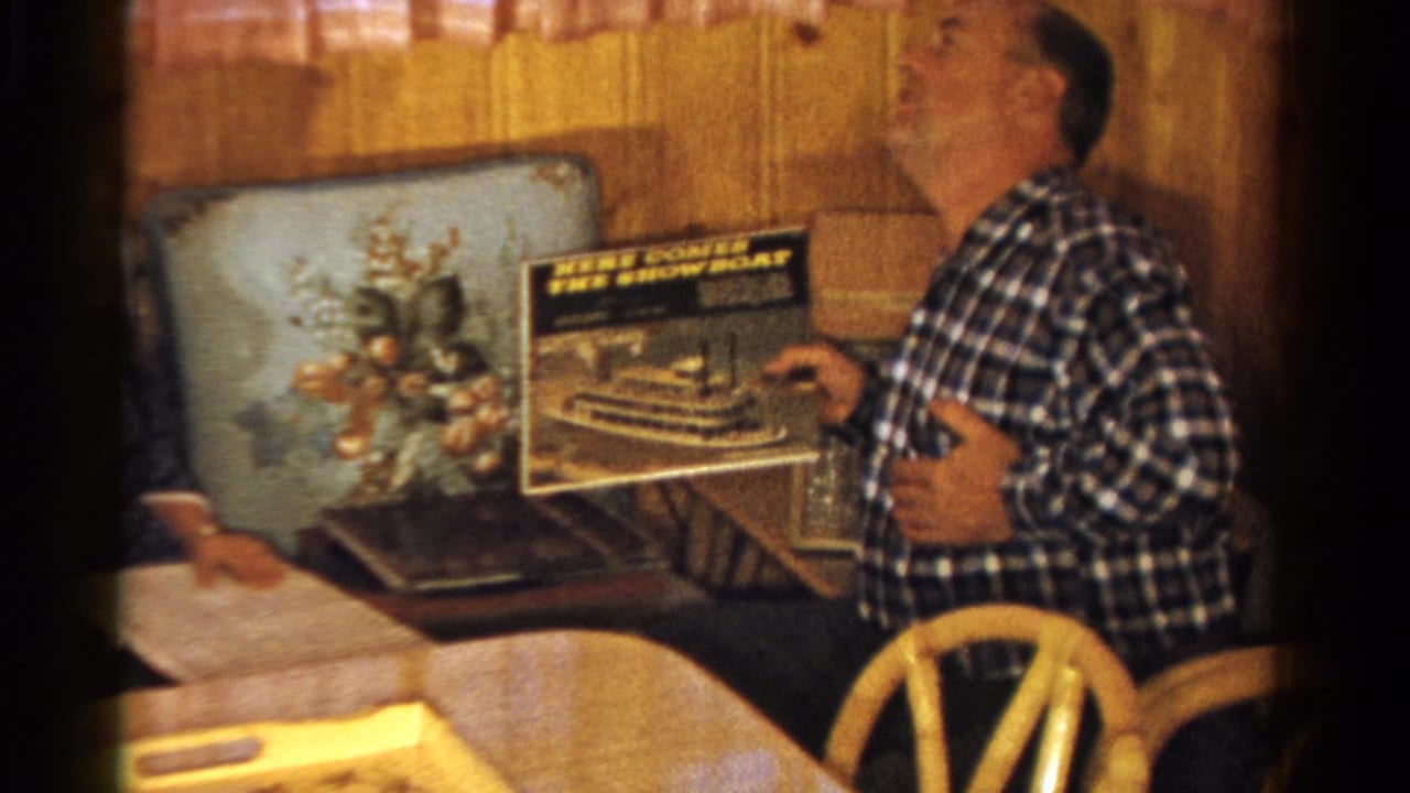 Two men sit in a wood-paneled room while one holds up a record album cover.