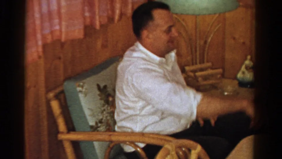 In the Ontario home interior, an adult man in a white shirt sits casually in a chair near floral upholstery and a green lampshade.