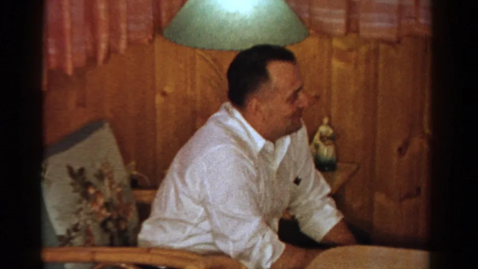 A 1958 Ontario home-movie frame shows an adult man in a white shirt leaning forward near a table under a green lampshade.
