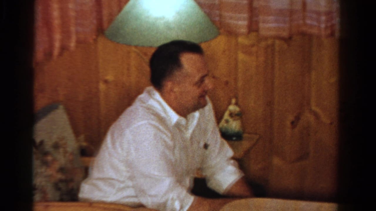 Adult man smiles in a wood-paneled Ontario home interior with a shaded lamp, side table, and patterned curtains, 1958 metadata supported.