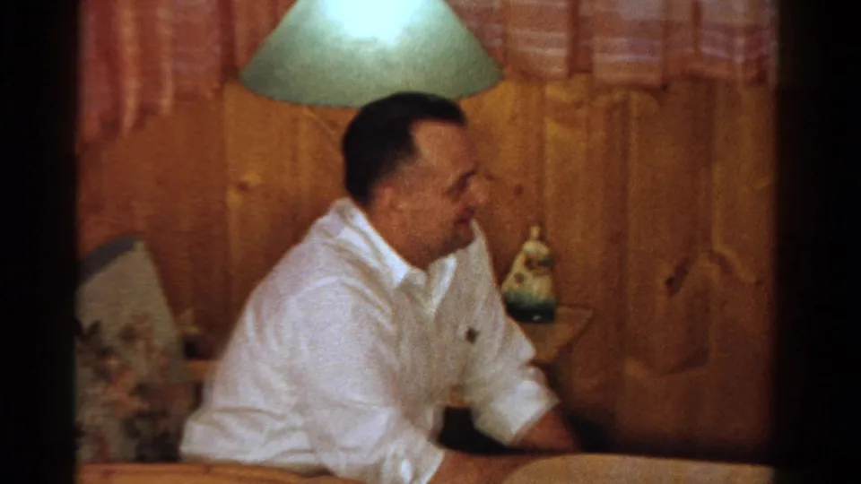 Adult man smiles in a wood-paneled Ontario home interior with a shaded lamp, side table, and patterned curtains, 1958 metadata supported.