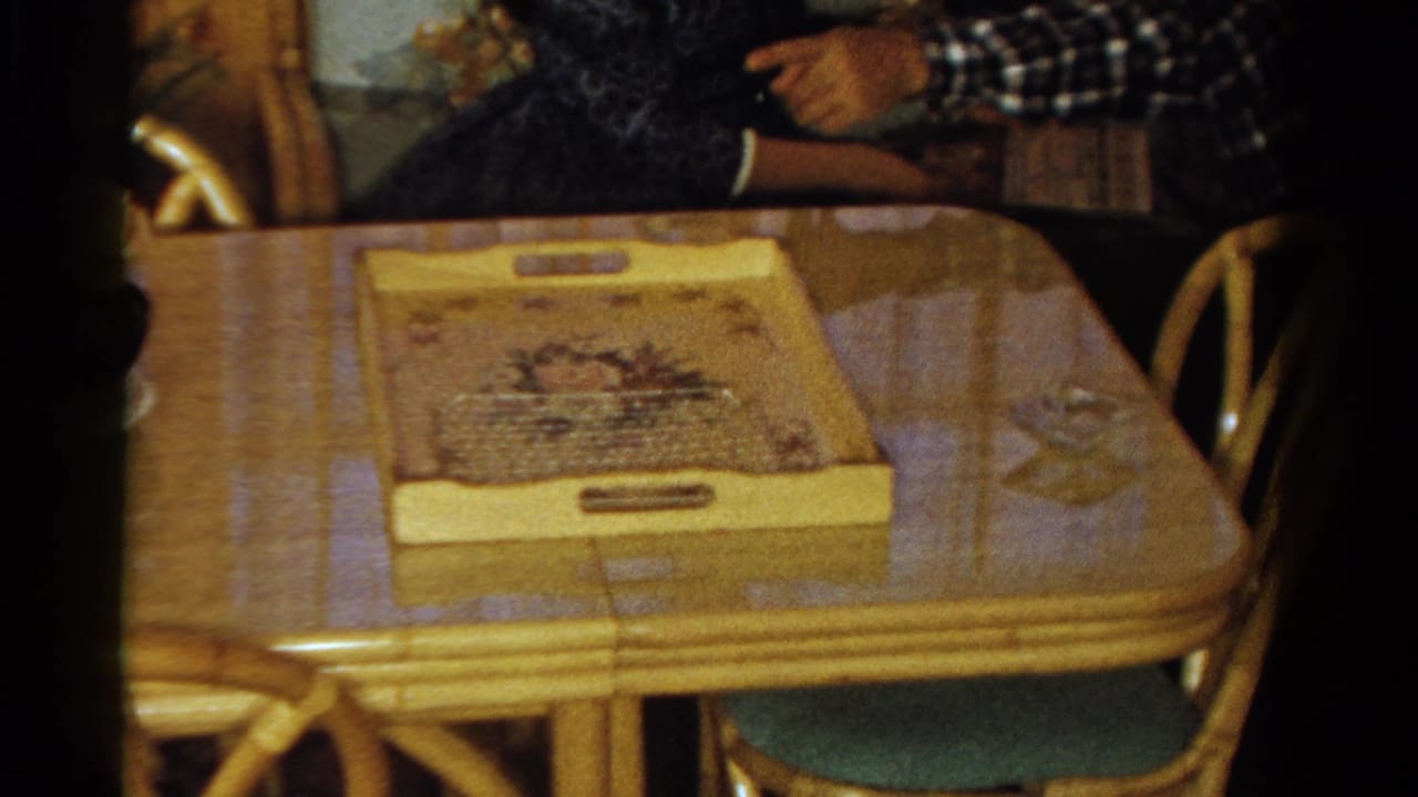 1958 Ontario home-movie frame with a decorative tray on a wooden dining table and an adult in a plaid shirt gesturing nearby.