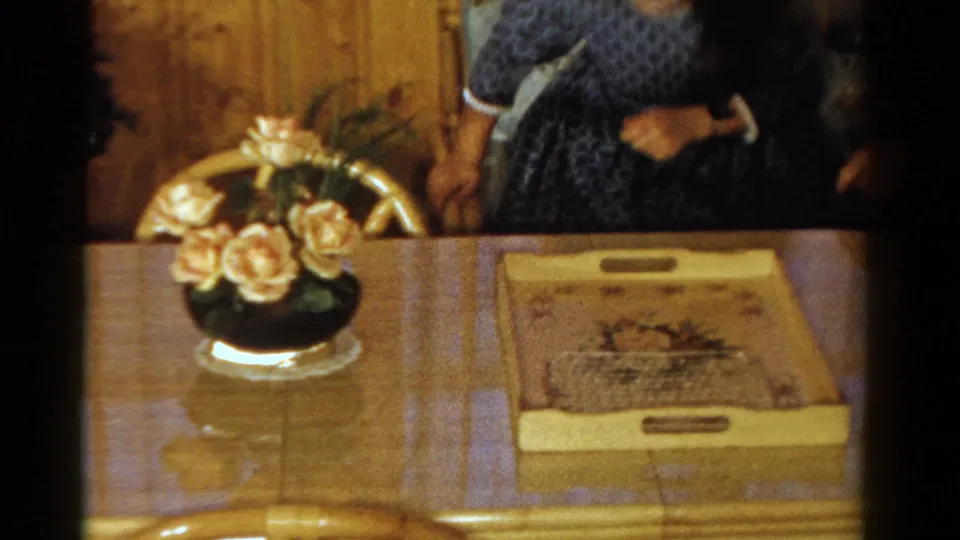 1958 Ontario home-movie frame showing an adult in a blue patterned dress beside a dining table with flowers, tray, and chairs.