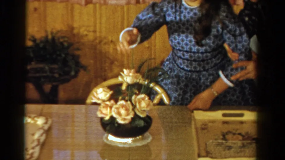 1958 Ontario home-movie frame showing an adult in a blue patterned dress seated near a flower arrangement and tray on a dining table.