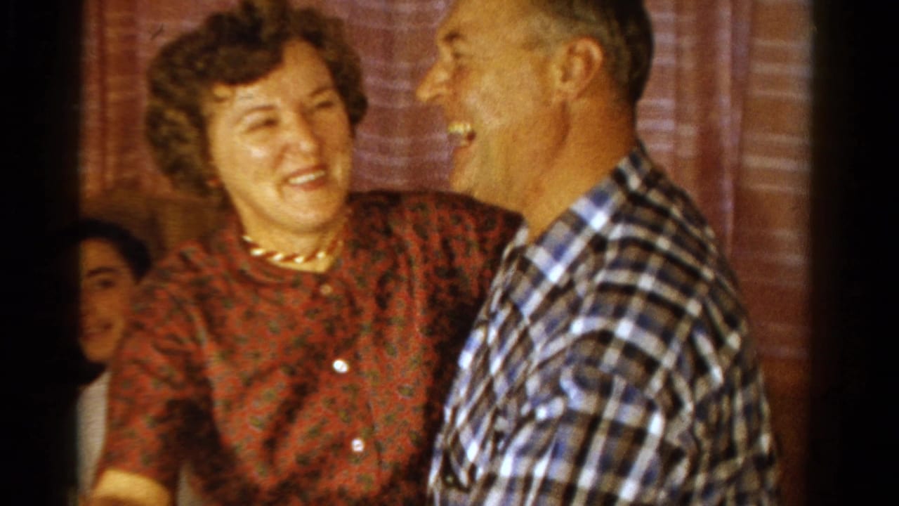 Smiling woman in a red patterned dress dances with a man in a plaid shirt indoors.