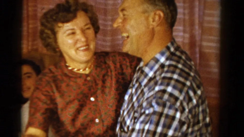 In 1958 Ontario home-movie footage, a woman in a red patterned dress smiles while dancing closely with a man in a plaid shirt.