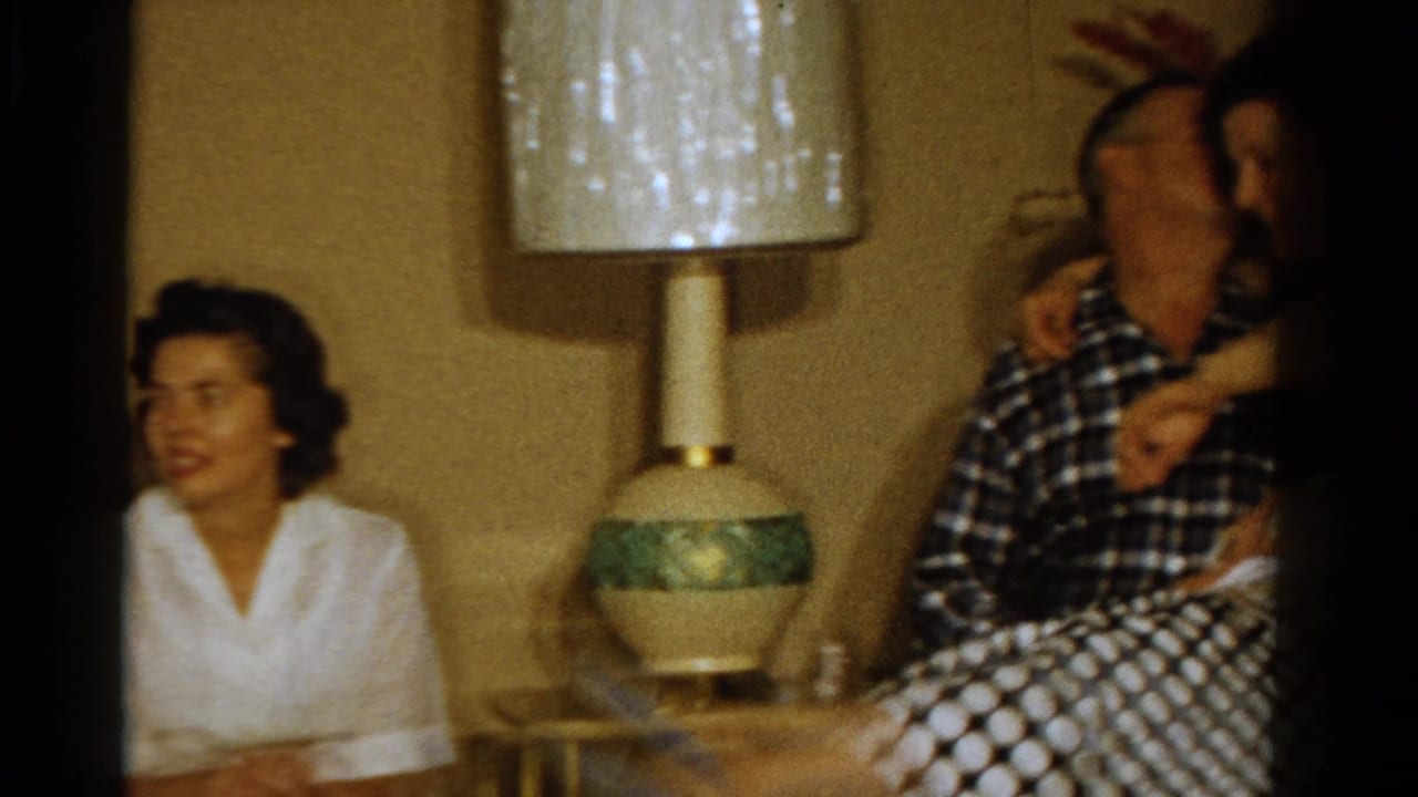 Ontario 1958 home-movie frame showing a woman in a white blouse and a seated couple near a textured table lamp.
