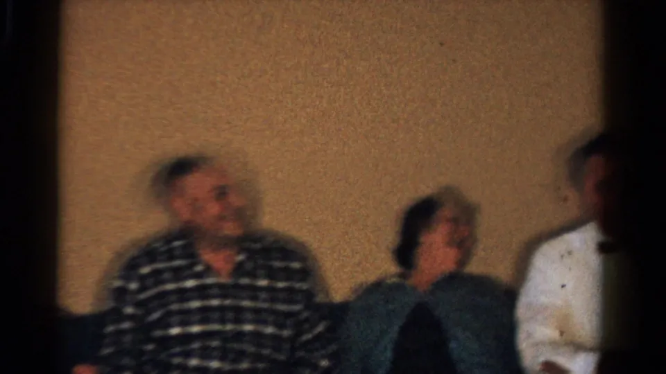 1958 Ontario home-movie frame showing seated adults in a warm interior, with a smiling man in a plaid shirt and others partly blurred by motion.