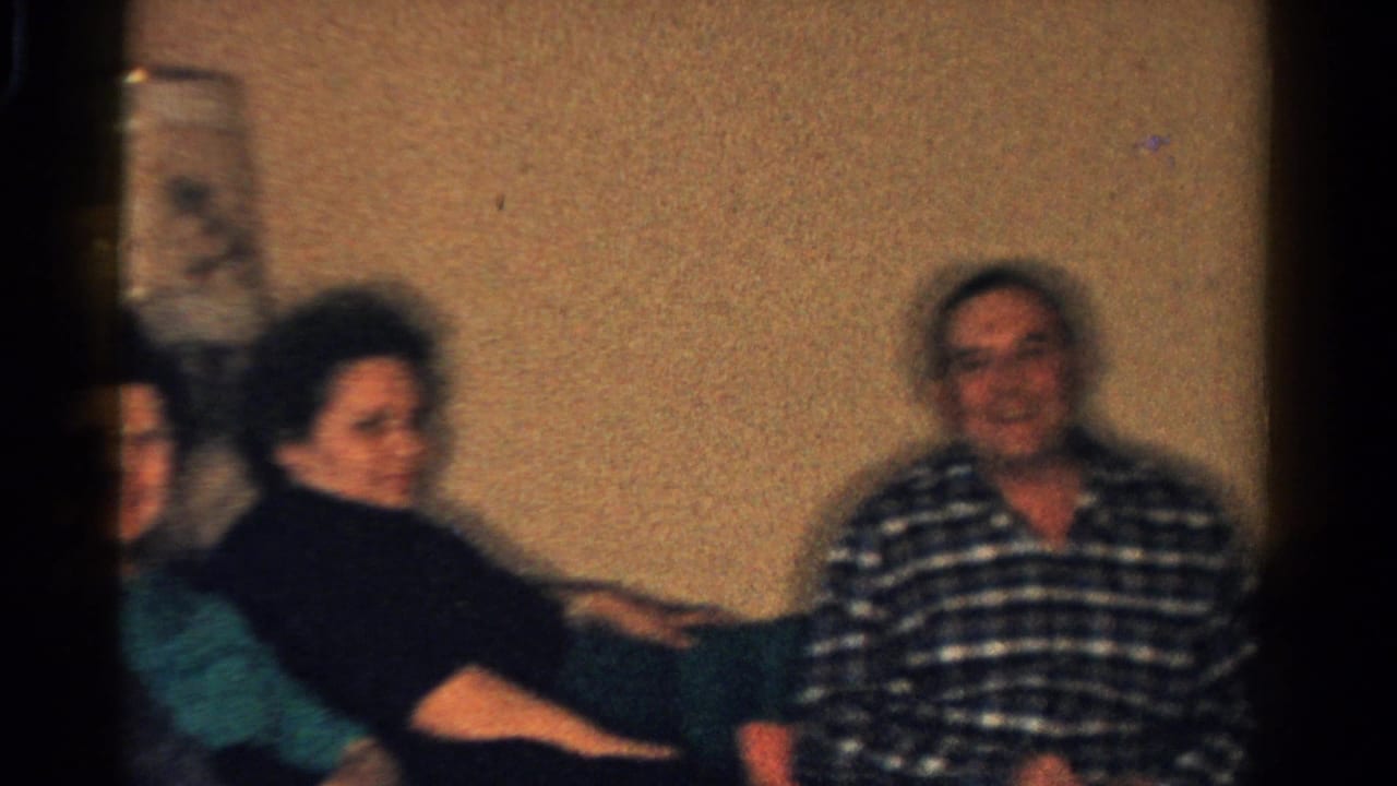 1958 Ontario interior frame with adults seated close together, including a woman in dark clothing and a smiling man in a plaid shirt.