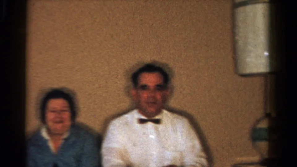 1958 Ontario home-movie frame showing a seated woman and a man in a white shirt with bow tie beneath a wall lamp.