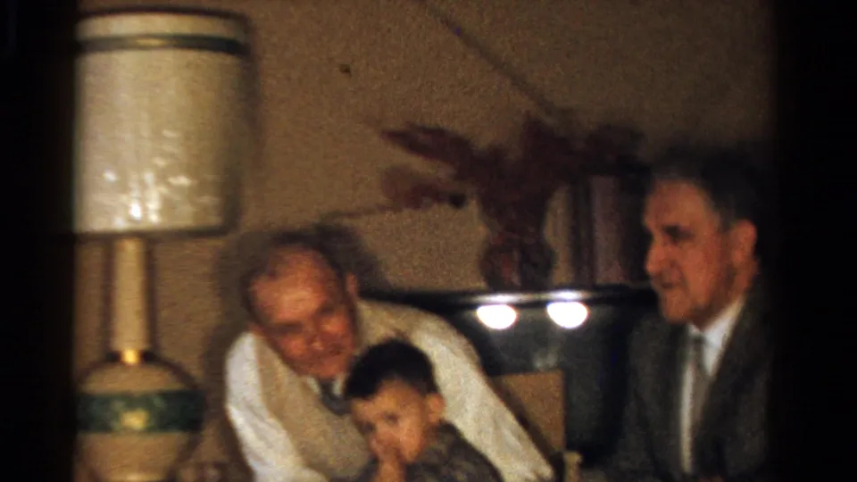1958 Ontario interior frame showing older adults and a young boy seated near lamps and a flower arrangement in a private room.