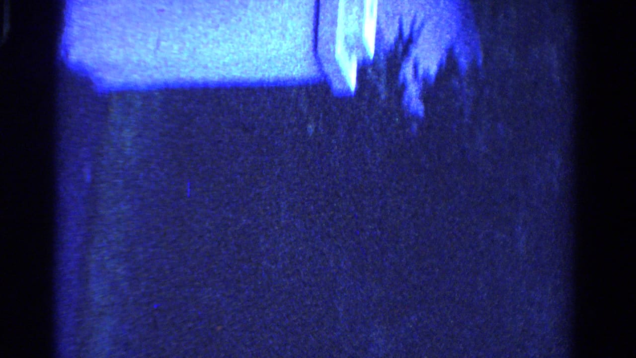 Blue-shifted 1958 Ontario 8mm frame, rotated sideways, with dark pavement and a partial view of a woman near the edge.