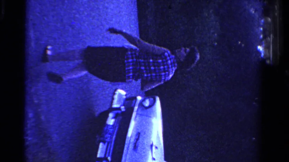 Sideways 1958 Ontario home-movie frame showing a woman walking past a light-colored vintage car on dark pavement.
