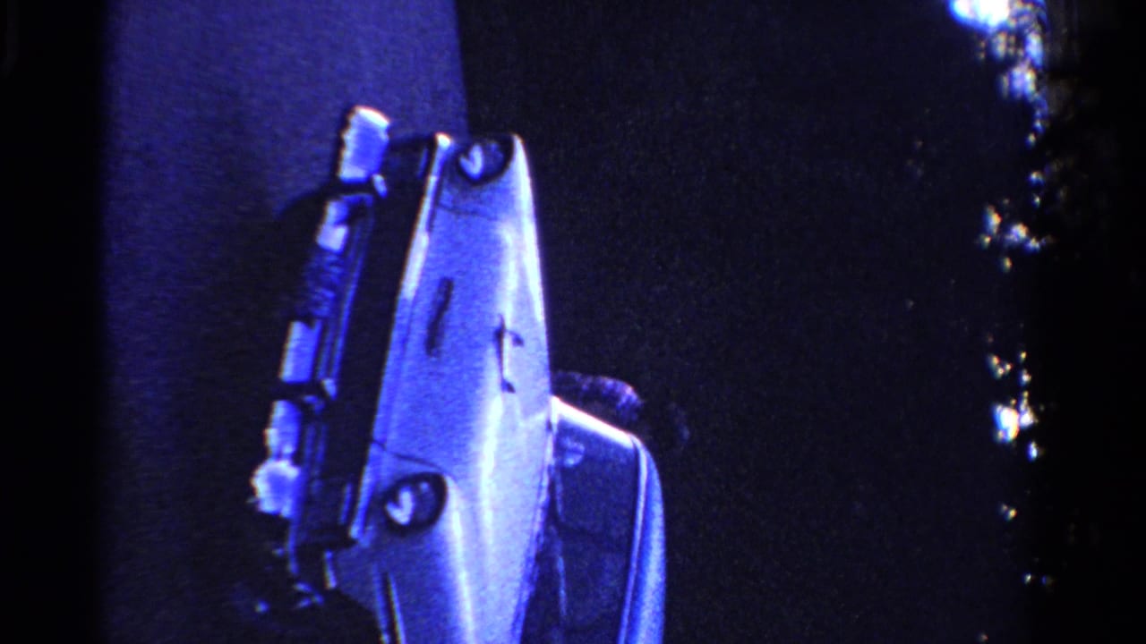 White sedan parked or angled beside a house wall in dark blue-tinted 8mm footage.
